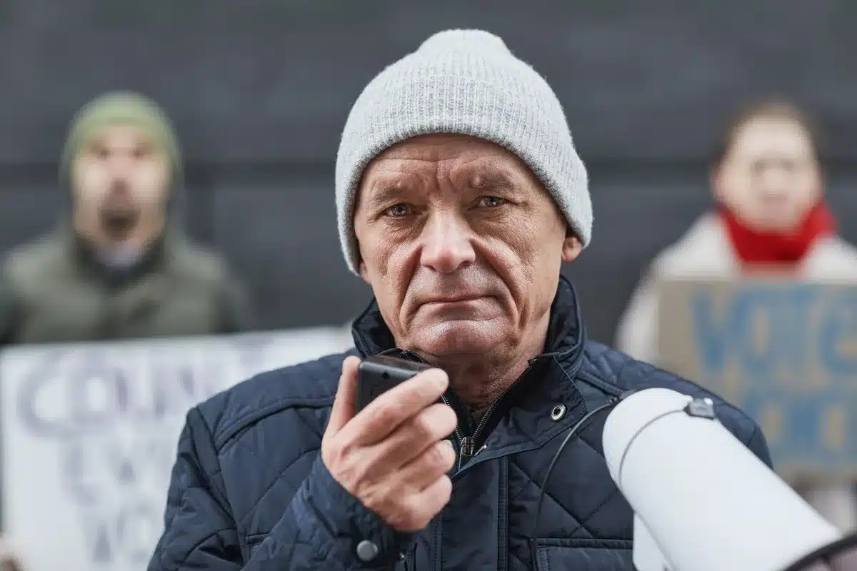 A politician at a campaign podium with supporters in the background at a rally, economic charts or housing imagery visible, showing campaign messaging shift toward affordability and economic issues.