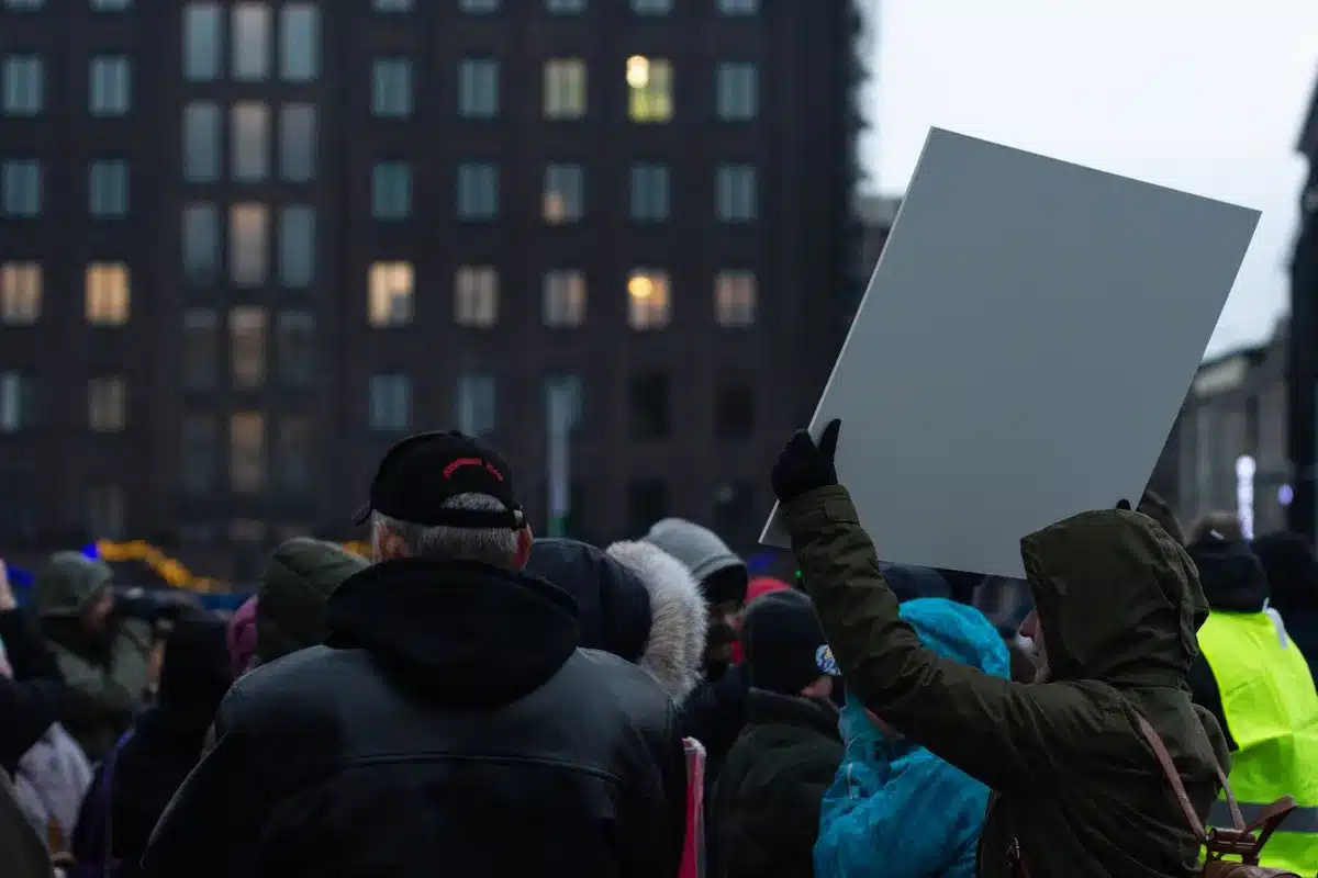 A group of figures in gray wizard robes and pointed hats holding a large banner with text, standing outside an ornate mansion gate at dusk, faces obscured by beards and shadows, a mix of determination and theatrical protest energy