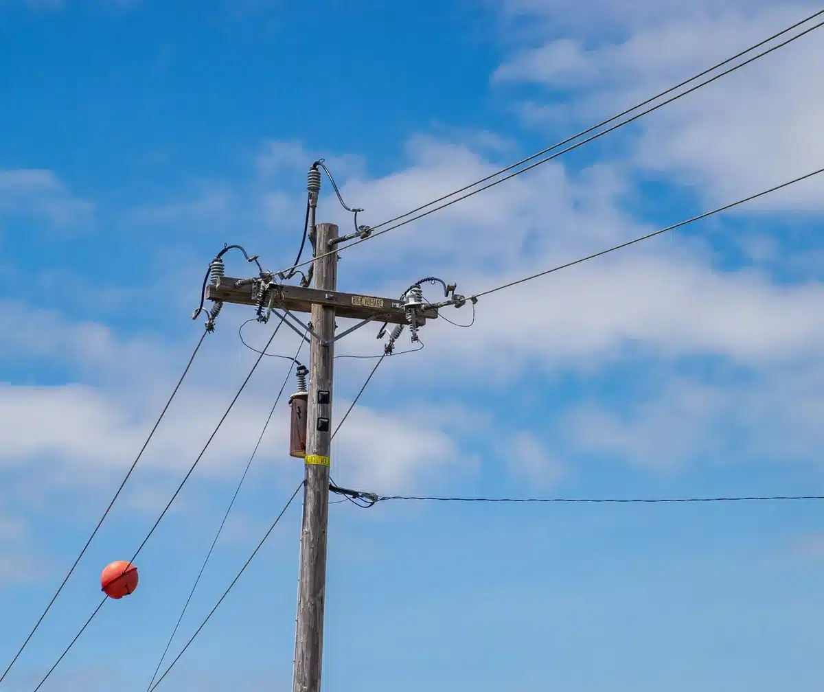 Close-up of aging electrical transformer equipment with rust and weathering visible, mounted on a utility pole against a gray sky, representing decades-old infrastructure near end of service life.