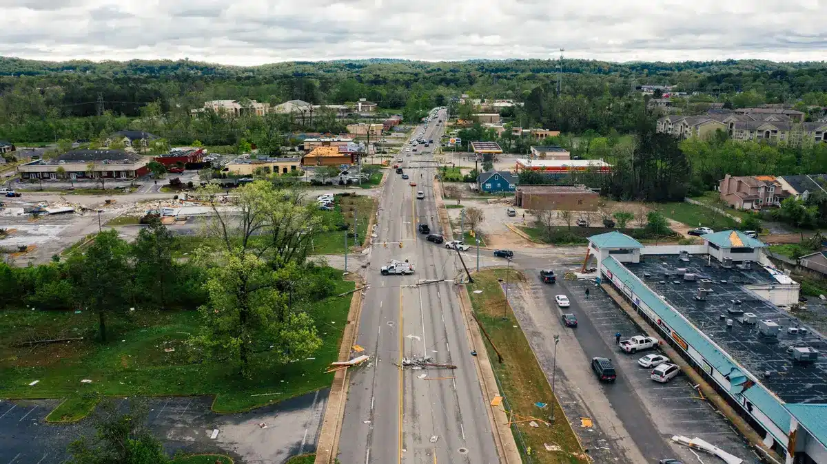 An empty residential street lined with downed trees and severed utility lines, power poles tilted at angles, debris scattered across pavement, overcast sky, no people visible