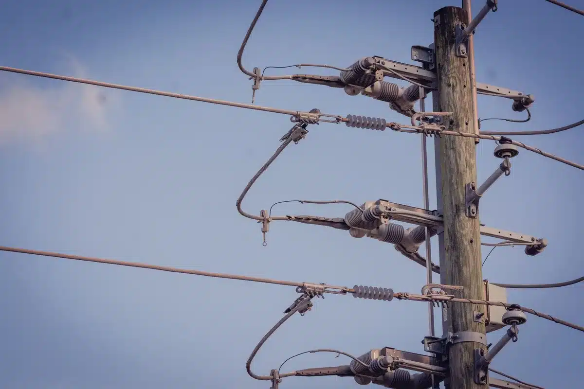 Repair crews working on damaged utility poles and downed power lines in daylight, with tools and equipment visible, emergency vehicles in the background