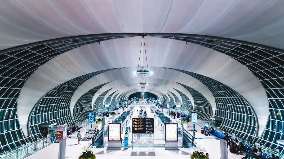 Modern airport terminal interior with departure boards, passengers with luggage, Qatar Airways signage and aircraft visible through windows