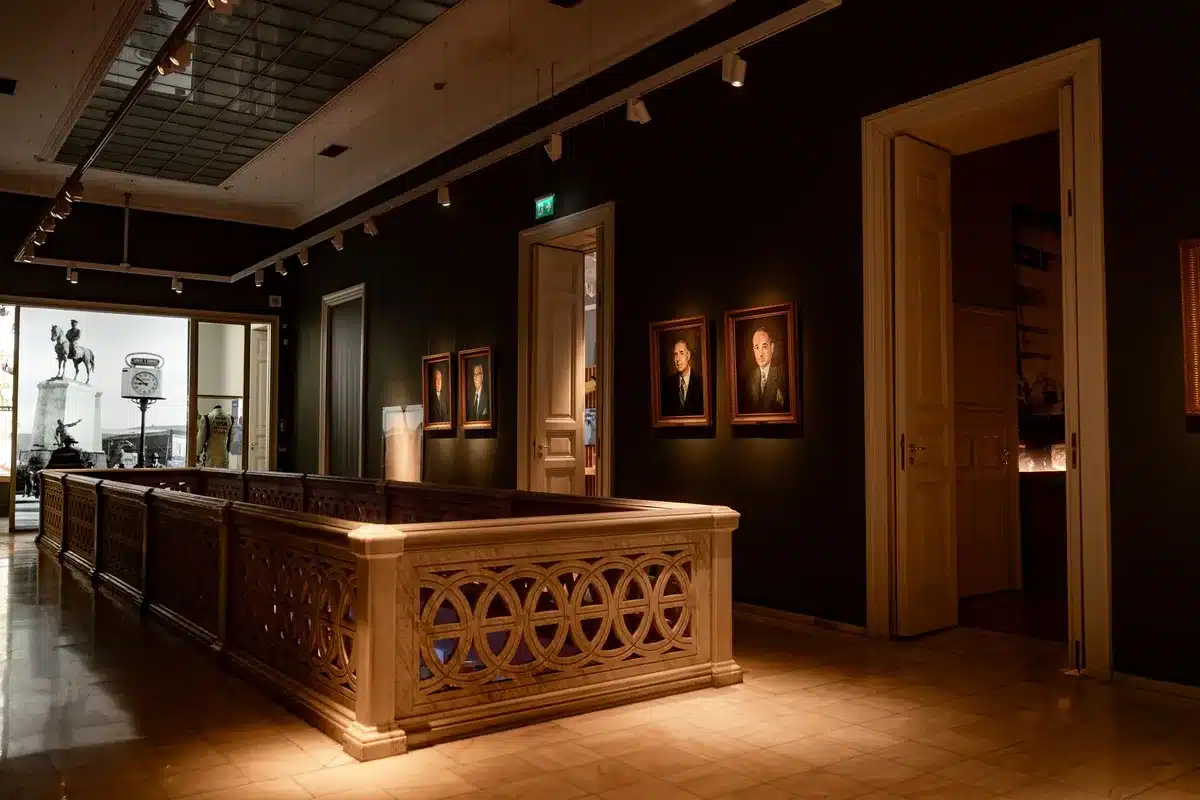 An elegant gallery space displaying formal royal gowns on mannequins under soft museum lighting, with ornate hats and accessories visible in glass cases nearby, visitors viewing the exhibits in the background