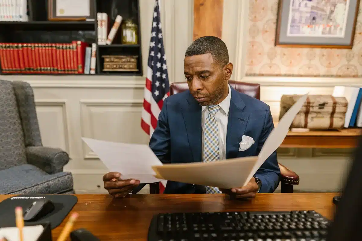 A formal government office setting with a desk, official seal, and empty chair facing toward a window; papers and a pen rest on the desk surface, suggesting administrative action and legal proceedings