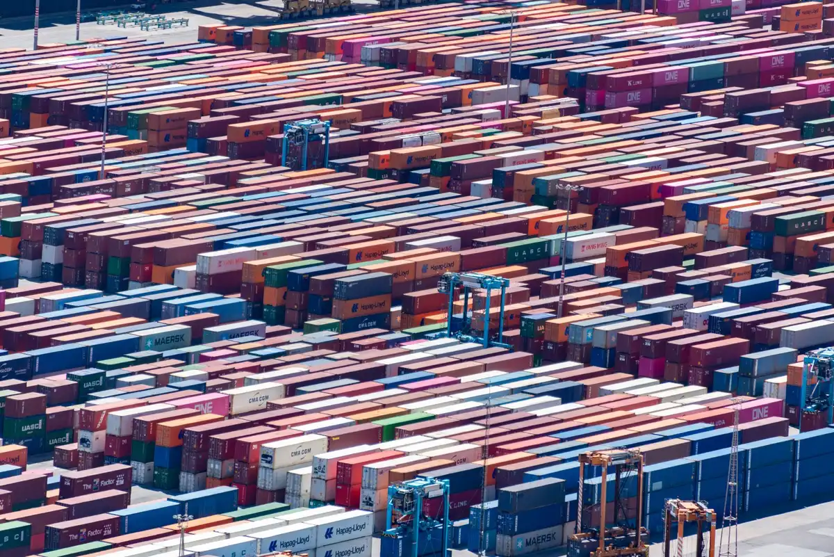 A split-screen view of an empty negotiation table on one side and stacks of shipping containers on the other, representing the breakdown in trade talks and escalating tariff barriers
