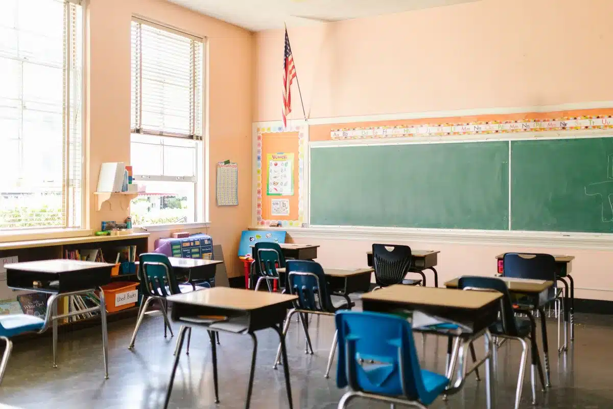 A row of empty classroom desks with vaccination pamphlets stacked on one desk, sunlight streaming through windows