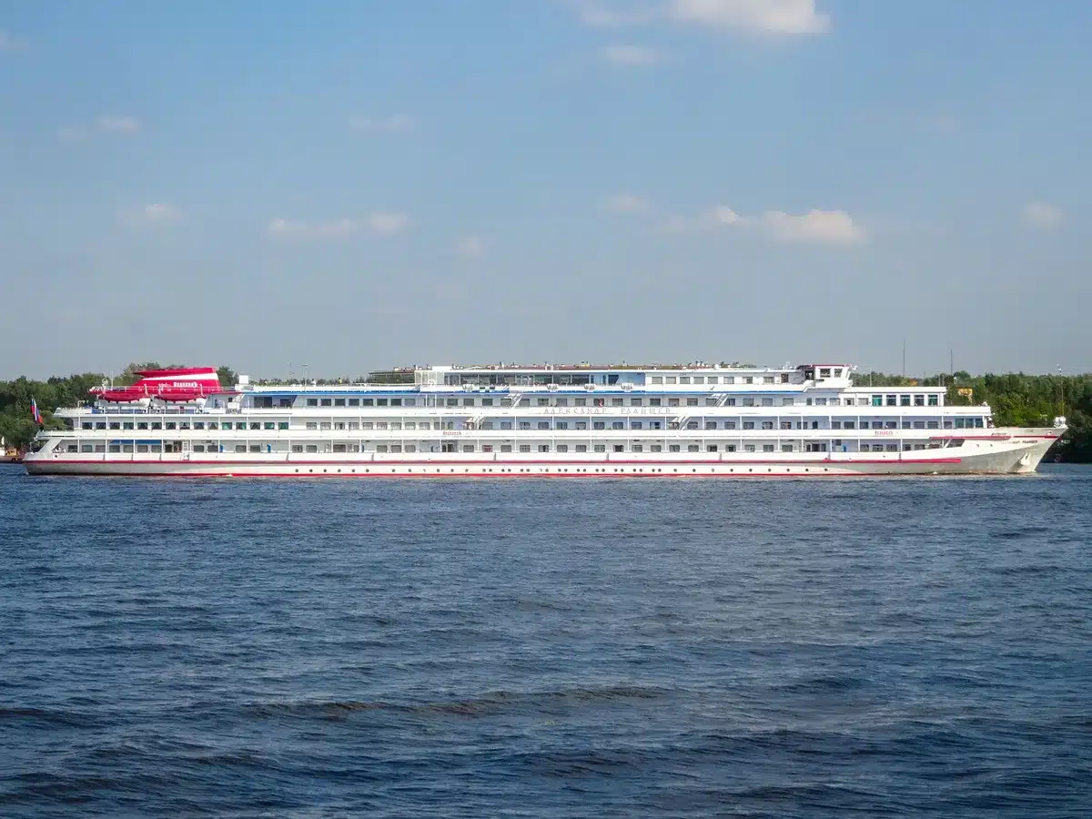 A cruise ship navigating shallow water with visible riverbed, crew working on deck adjustments, river landscape with low water line marked on banks