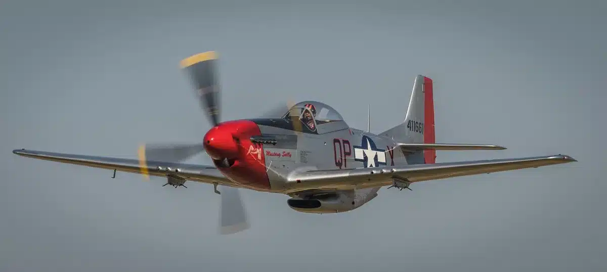 Historic P-51 Mustang aircraft on an airfield tarmac, propeller gleaming in sunlight, vintage military paint scheme visible, ground crew nearby, blue sky overhead