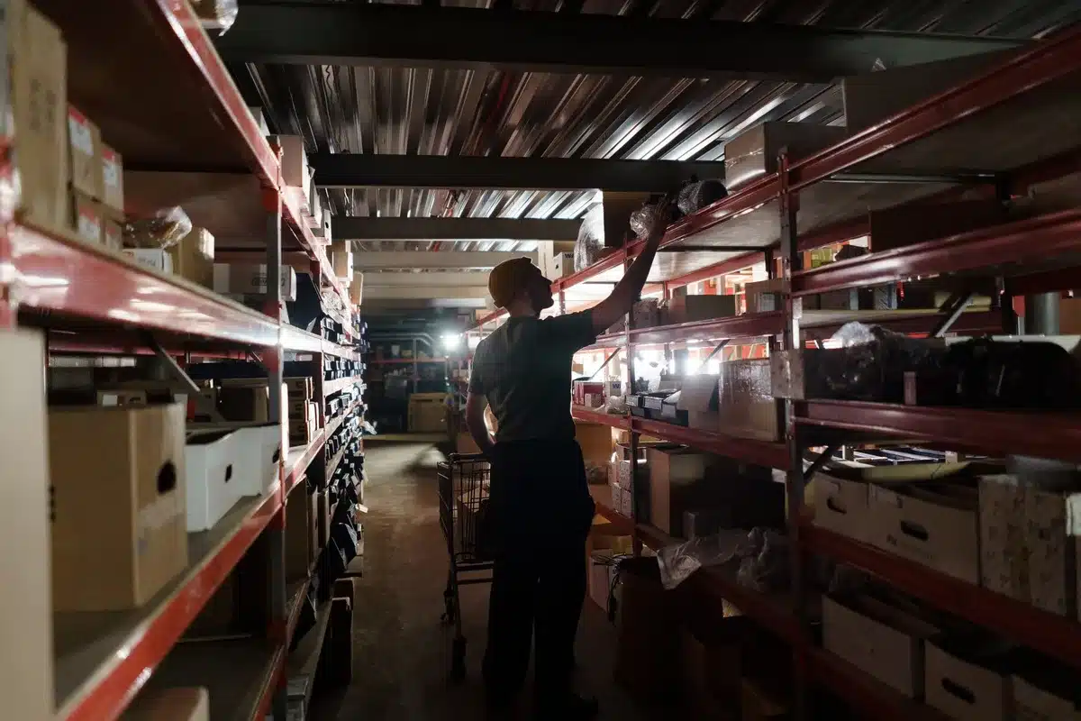 A modern distribution center interior with neatly organized inventory shelves, merchandise boxes stacked efficiently, warehouse staff moving through the space, bright industrial lighting overhead, conveyor systems in motion.