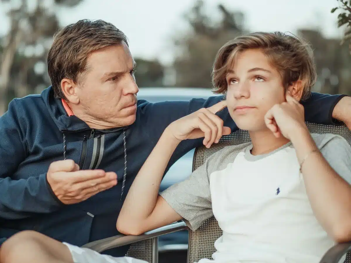 A parent and child sitting in a car, the child gazing out the window while the parent drives, morning light streaming through the windshield, a smartphone on the dashboard glowing faintly.