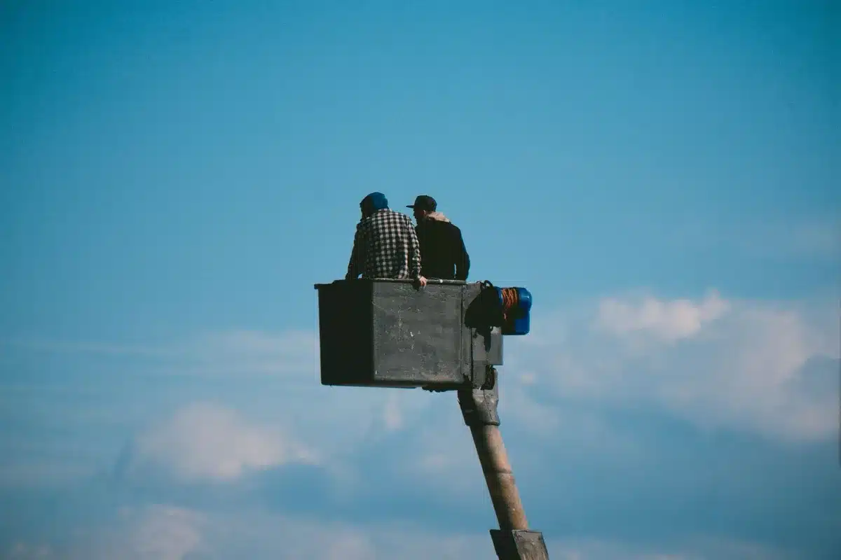 A crane truck bucket elevated near a streetlight under a highway overpass, the bucket empty, a damaged box truck visible below, daylight, urban infrastructure setting
