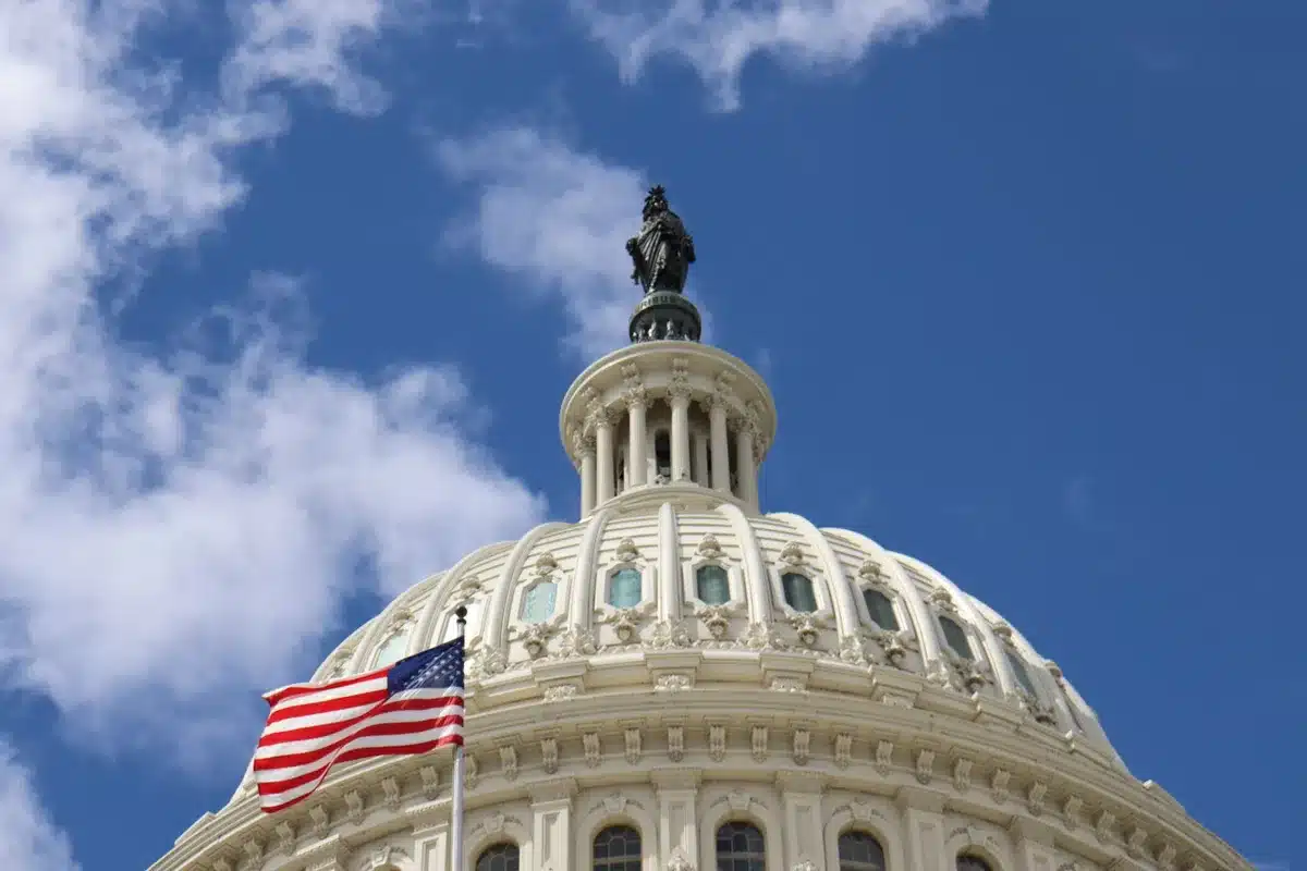 A modern state capitol building exterior with columns and steps, American flags flanking the entrance, overcast sky