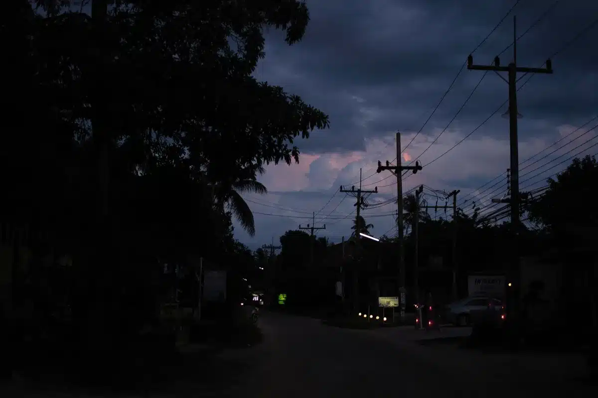 Darkened residential street at dusk with streetlights off, utility poles silhouetted against dimming sky, a single emergency vehicle light visible in distance