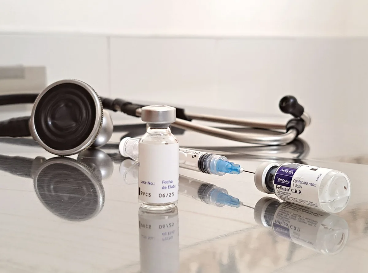 A pediatrician's office vaccination station with syringes, vaccine vials, and immunization records on a desk, morning light through a window, the clinical setting calm and orderly, trust and care implicit in the preparation.