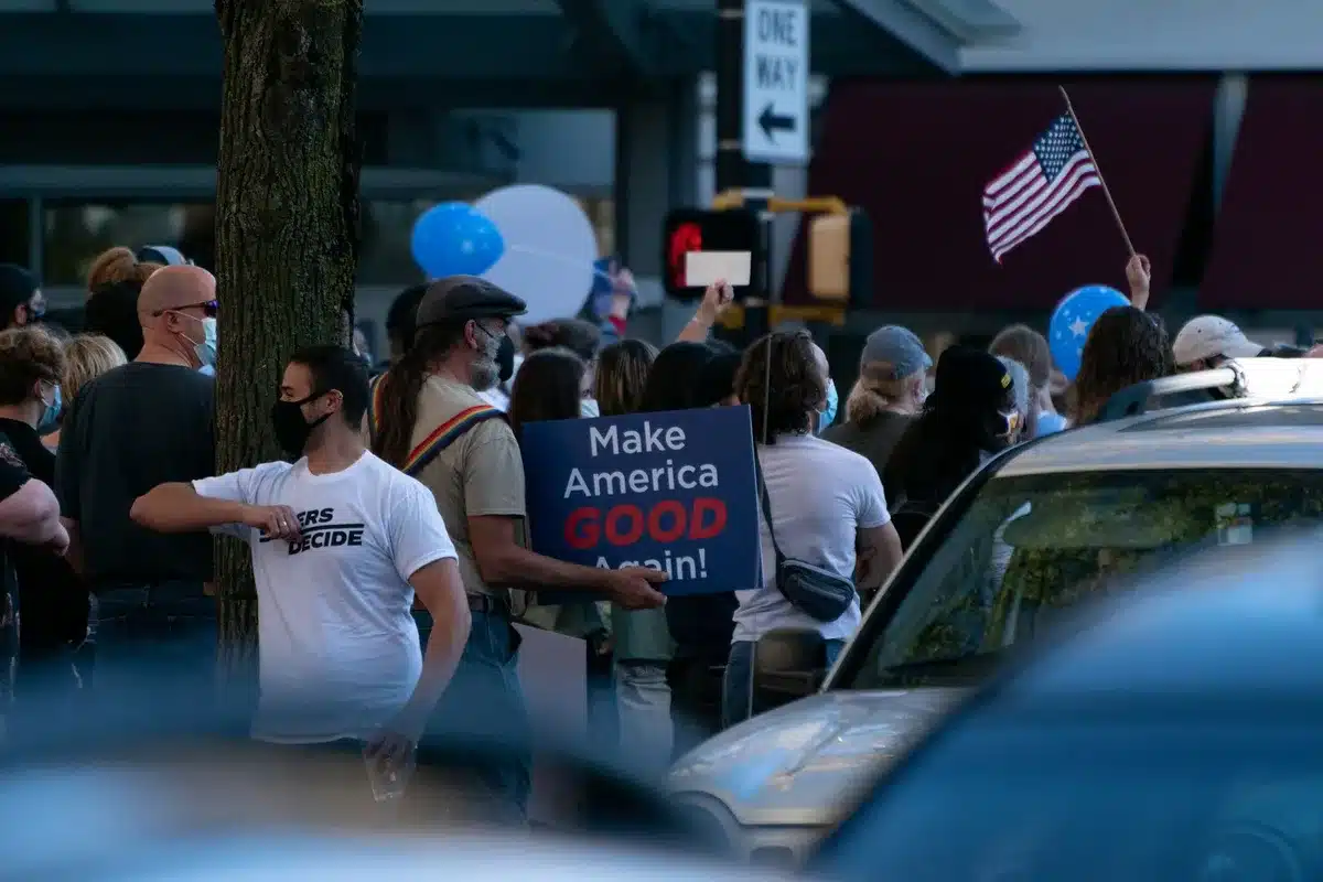 A crowded rally venue in Toledo with supporters holding campaign signs, diverse attendees with raised arms, faces animated with engagement, stage lighting and blue-and-red campaign colors visible, autumn afternoon light streaming through windows