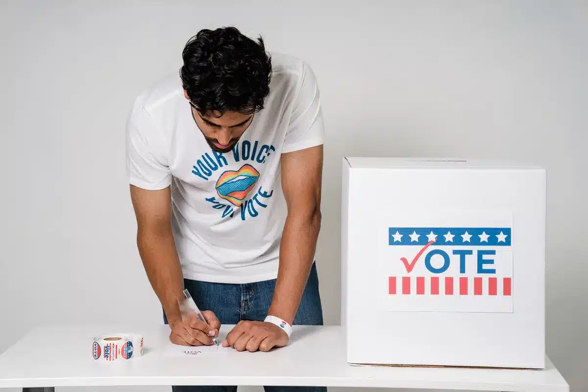 A close-up of a ballot box with a security seal, American flags in soft focus behind it, natural lighting from a window, emphasizing the vulnerability of election materials.