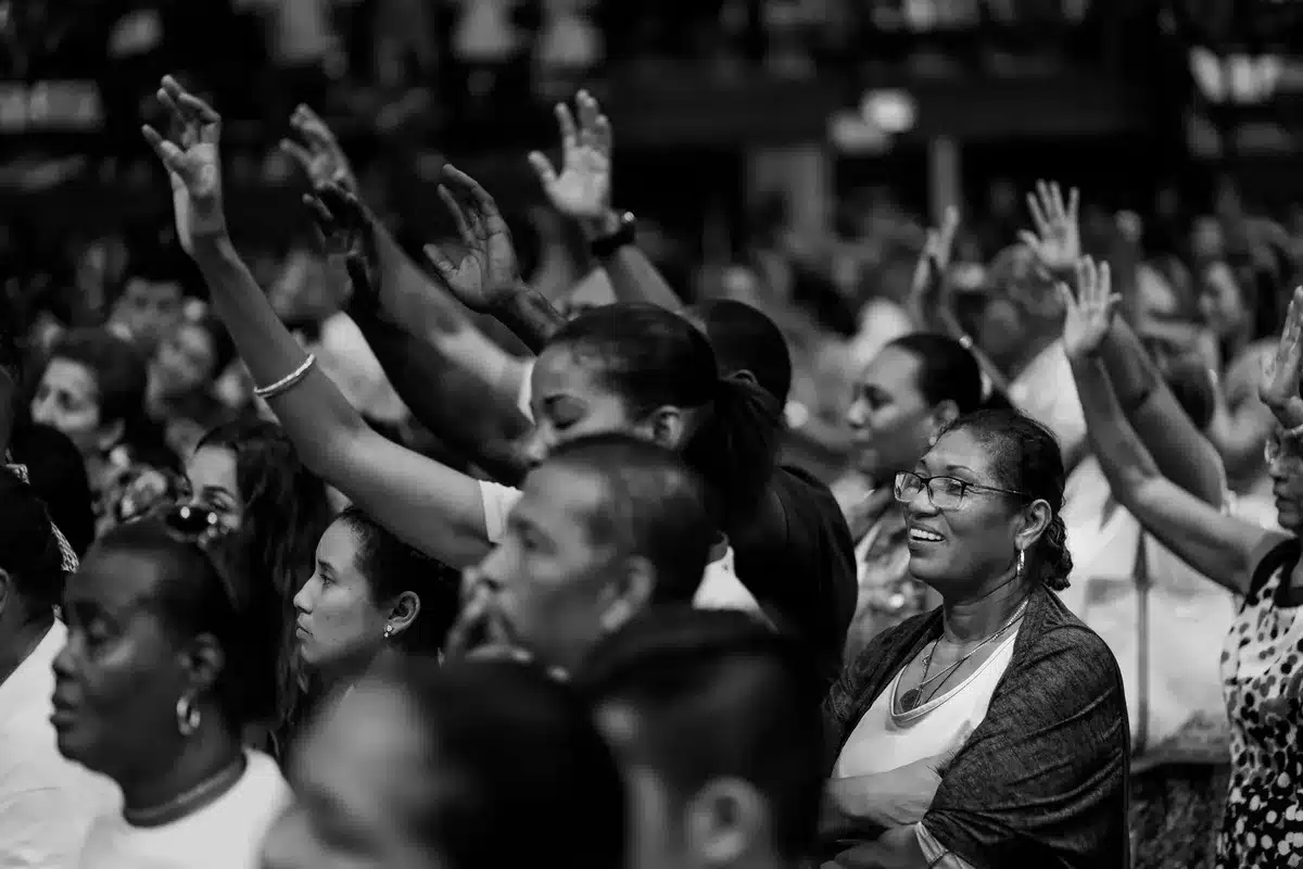 A crowd of anonymous supporters in stadium seating, some standing with raised arms, faces blurred or distant, watching a stage below, warm stage lighting casting long shadows, collective anticipation visible