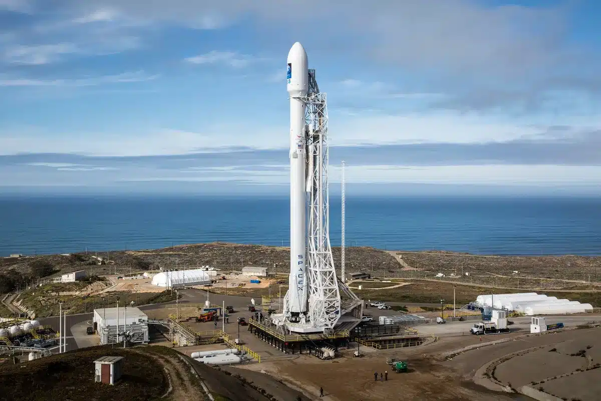 A Falcon 9 rocket ascending at dawn from a coastal launch pad, exhaust plume illuminated by early light, booster flame bright against the dark sky, the Pacific Ocean visible in the distance, power and routine converging