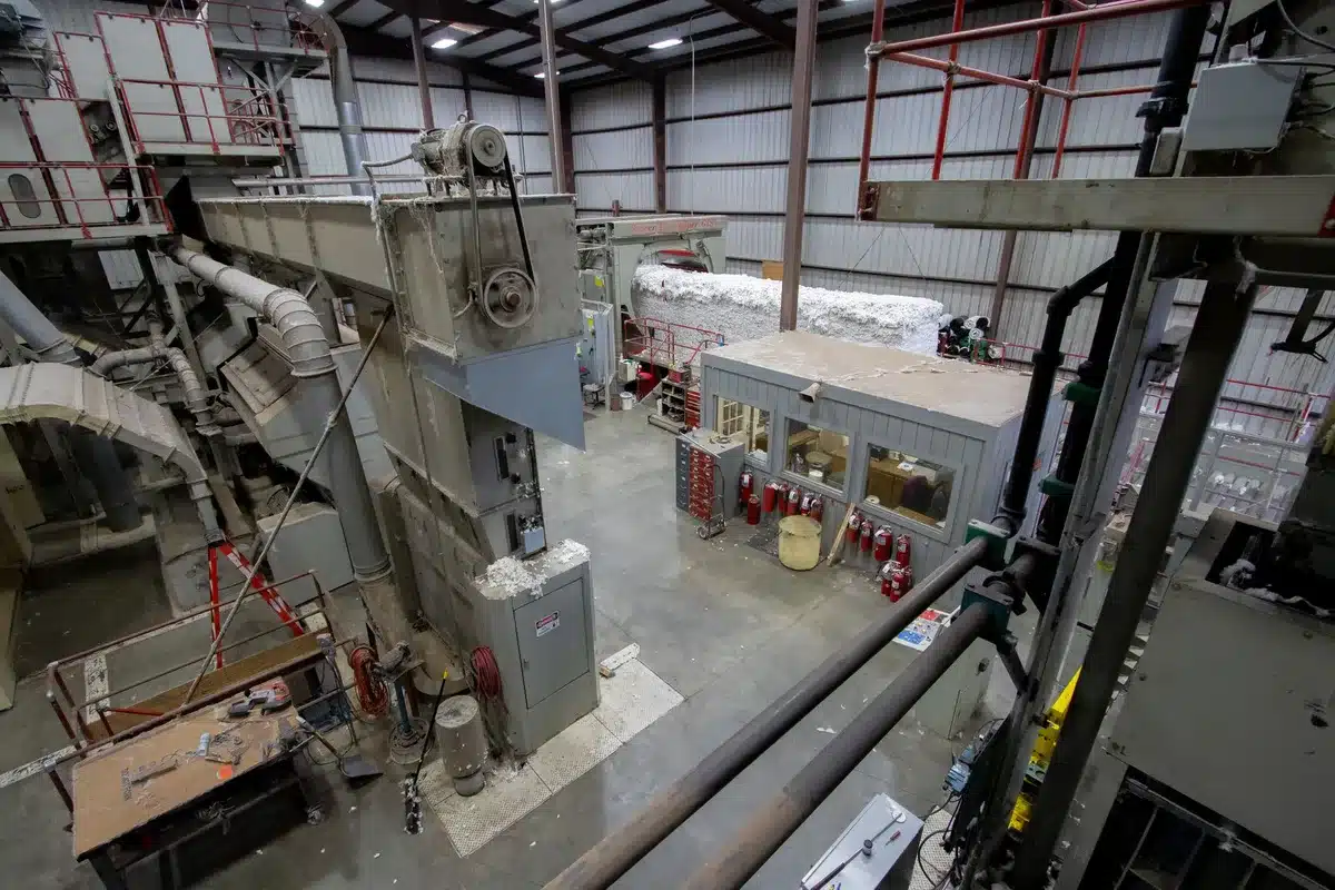 Interior of an aerospace manufacturing facility, a Falcon 9 second stage under bright work lights on an assembly platform, technicians in the background checking systems, partially-assembled rockets visible in the hangar, the scale and precision of production evident