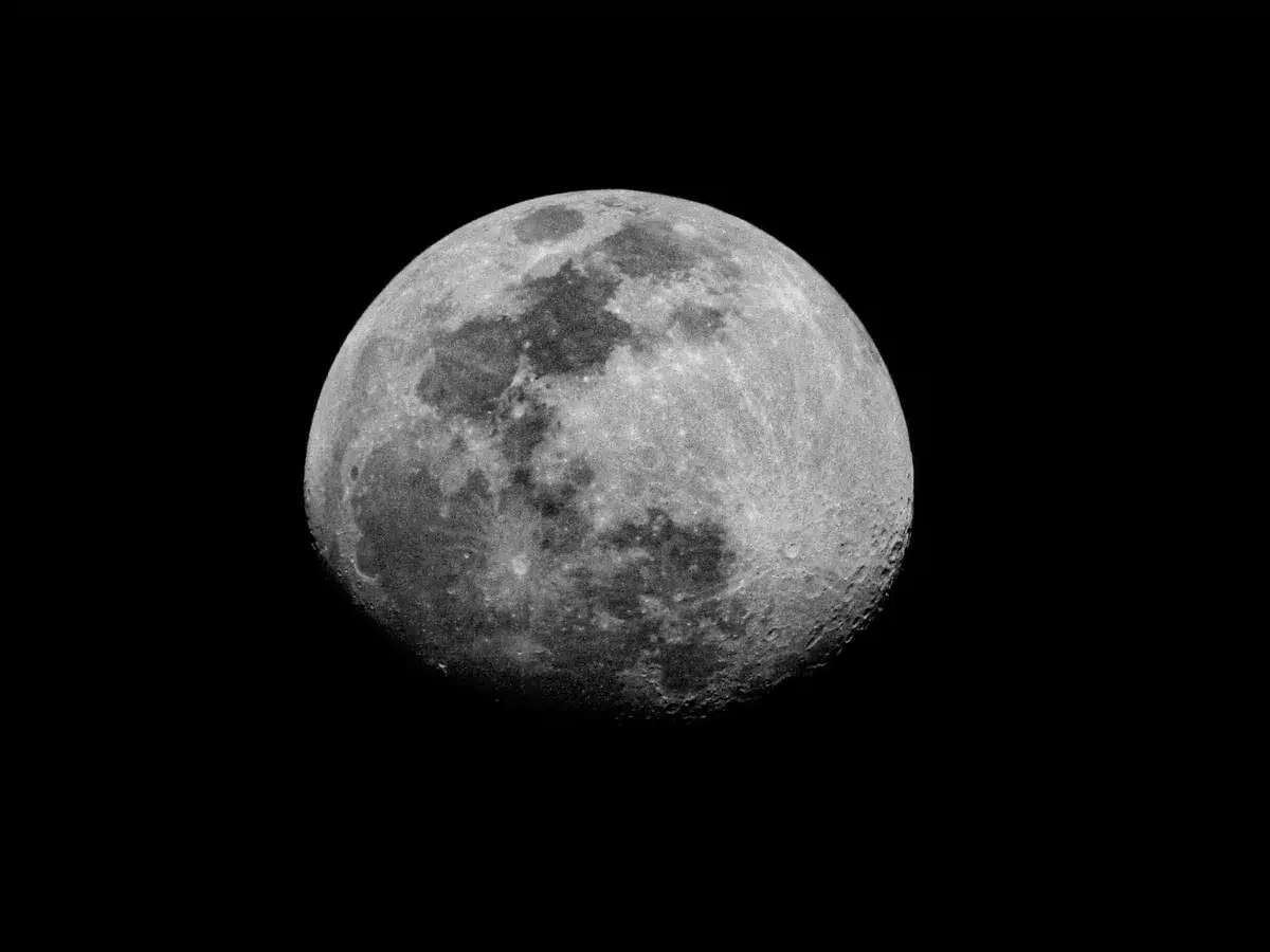 A barren lunar surface with a fresh impact crater surrounded by ejected dust and regolith, sunlit on the Moon's near side, showing the scale of the newly carved depression and debris field.