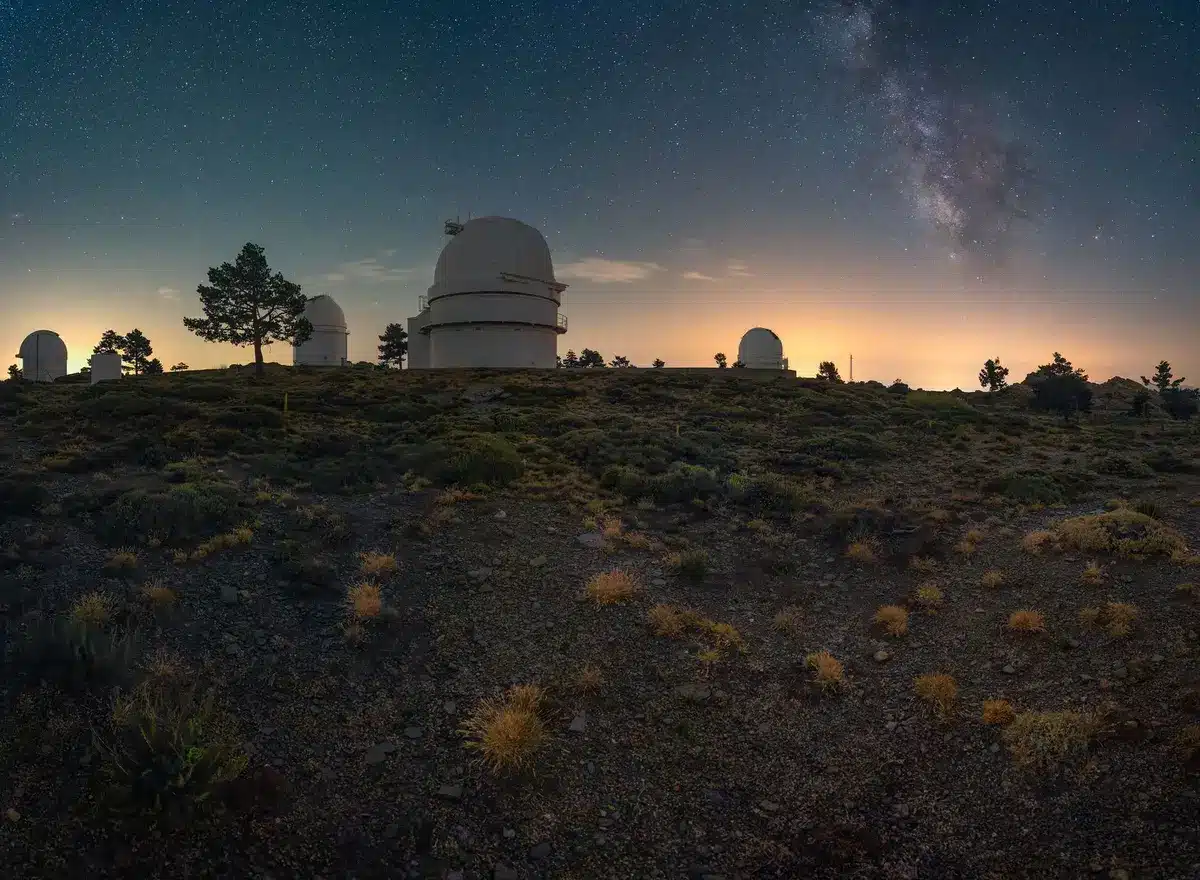 A telescope dome silhouetted against a starry night sky, with the Moon visible above the horizon, representing the observation of the impact event from Earth.
