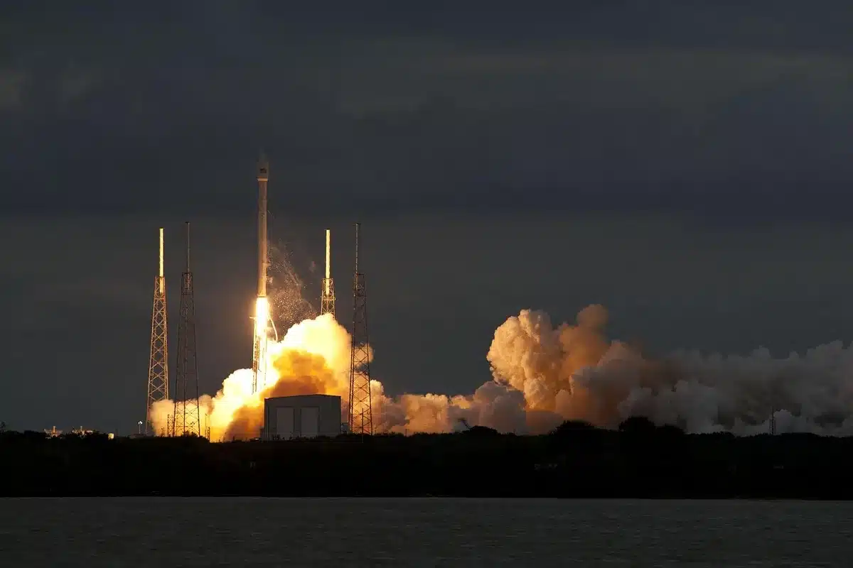 A Falcon 9 rocket on the launch pad at Vandenberg Space Force Base at dusk, orange and white rocket standing against darkening sky, launch tower lights illuminating the structure, the Pacific coast visible in the background