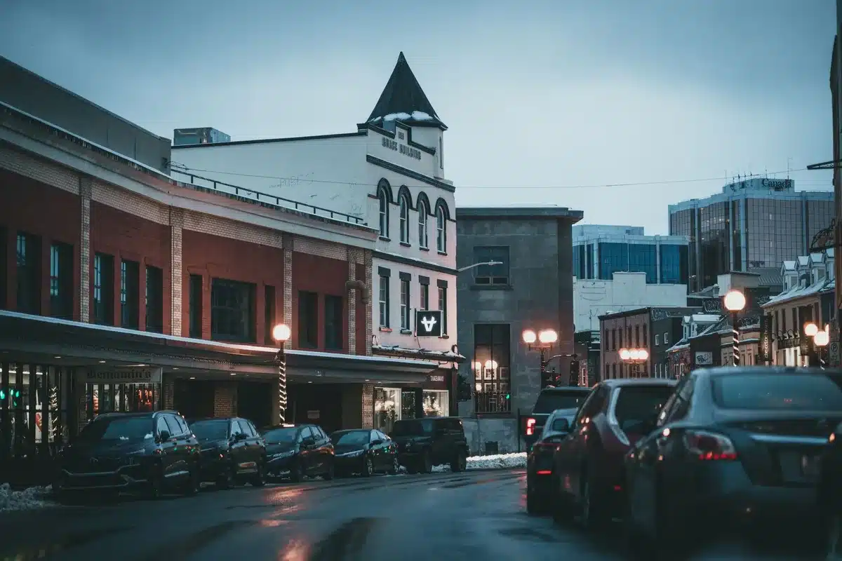 An empty parking lot at dusk with unlit streetlights and dark storefronts, a few distant car headlights, power lines silhouetted against a twilight sky