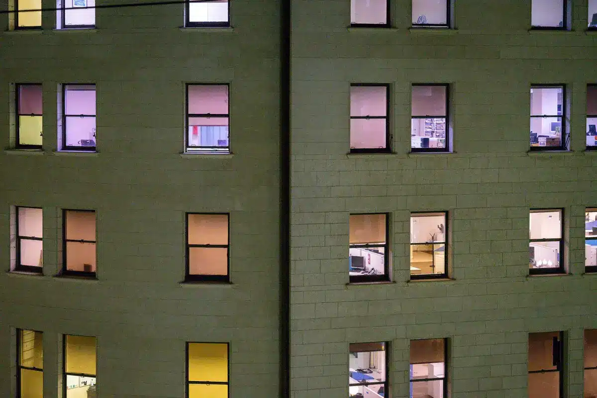 A corporate office building with modern glass windows, interior workspace visible, desks and workstations empty or sparsely populated, muted lighting