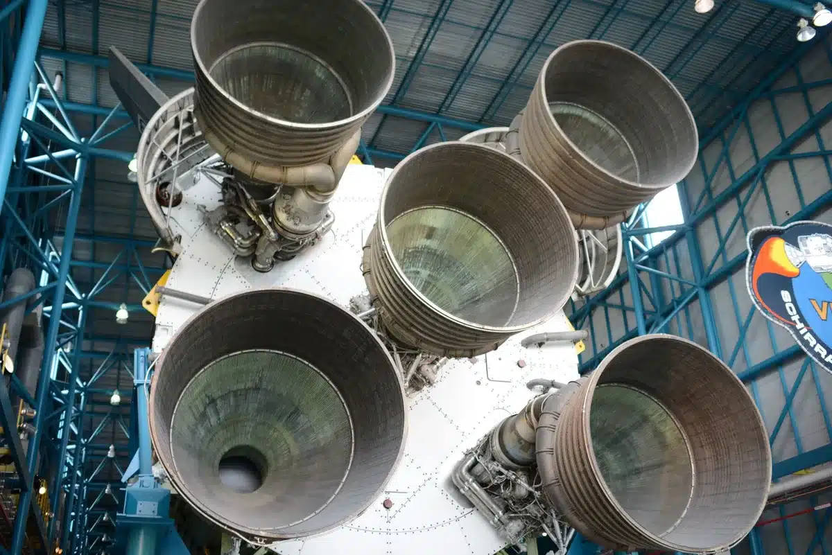 A stainless steel rocket upper stage section suspended in a dry dock facility, industrial cranes positioned overhead, workers in safety gear visible at a distance, suggesting preparation for refurbishment and reuse