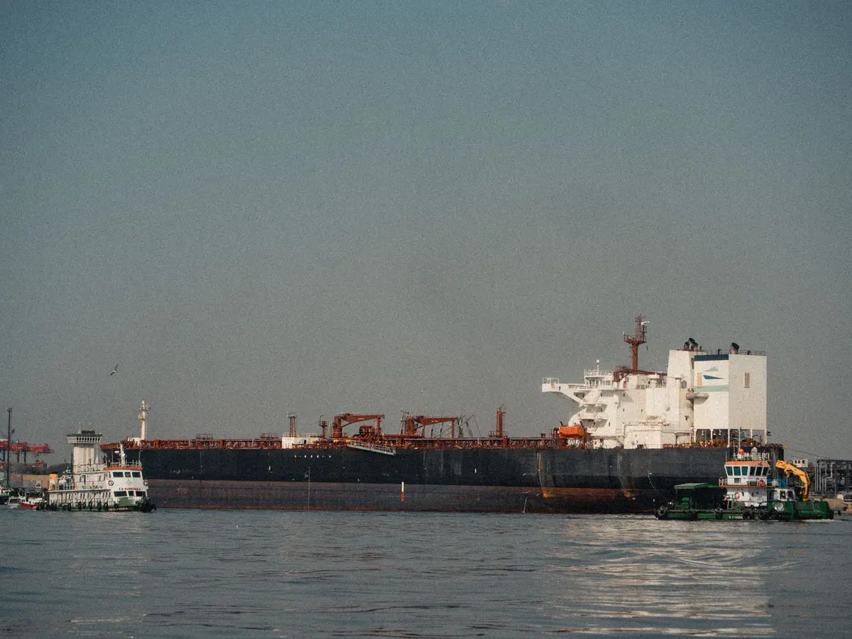A cargo ship navigating through narrow waterway with warning lights, surrounded by military vessels in the distance, representing the strategic chokepoint and geopolitical tension