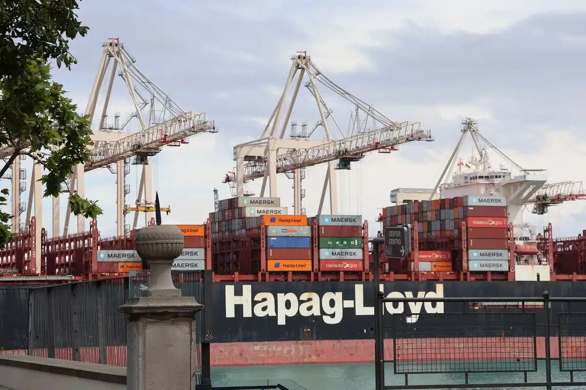 Container ships lined up at port terminal during dusk, industrial cranes silhouetted against sky, global trade infrastructure at standstill, empty berths visible, energy sector equipment