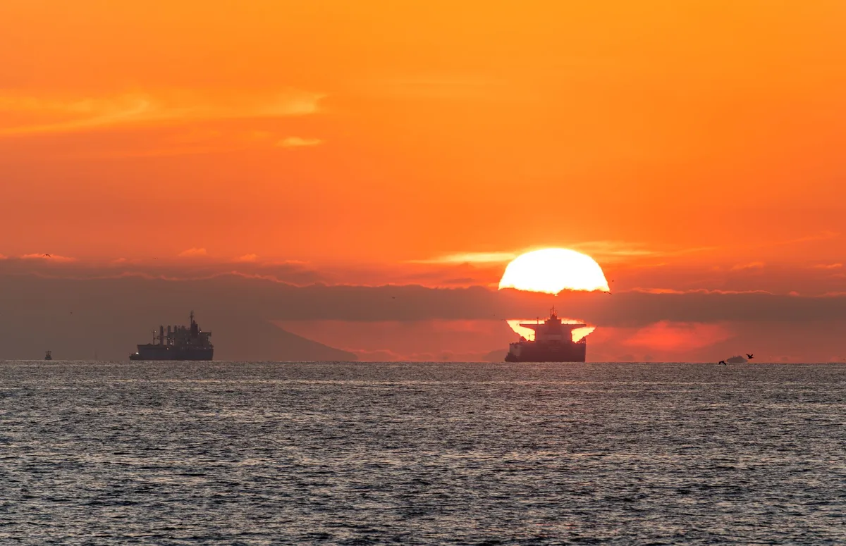Oil tanker ship silhouette against a darkening horizon, viewed from a distance, anchored in calm blue-grey water with industrial port structures blurred on the shoreline