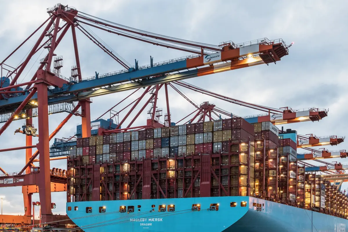 Empty commercial shipping container port at dusk, berths standing vacant with no cargo vessels, cranes silent against a grey sky, one distant ship visible on the horizon