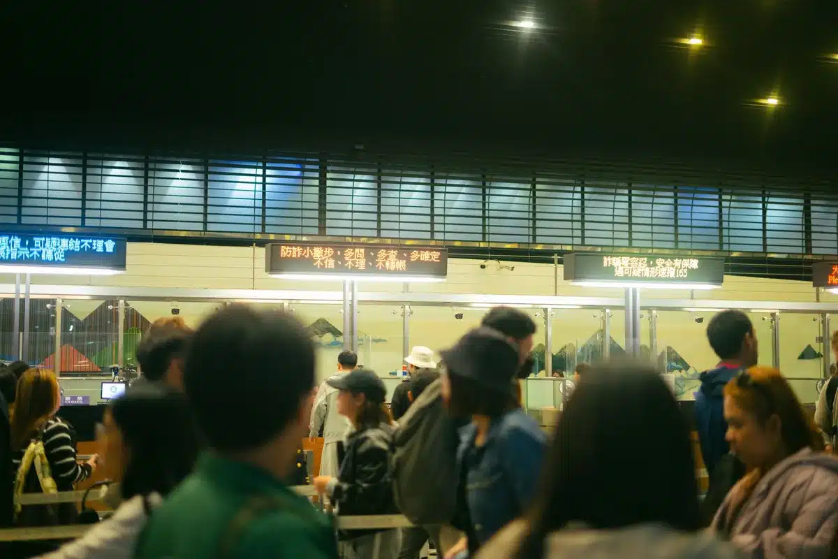 A crowded airport security checkpoint viewed from above, with anonymous travelers in motion blur standing in line, fluorescent lighting casting harsh shadows on the floor.