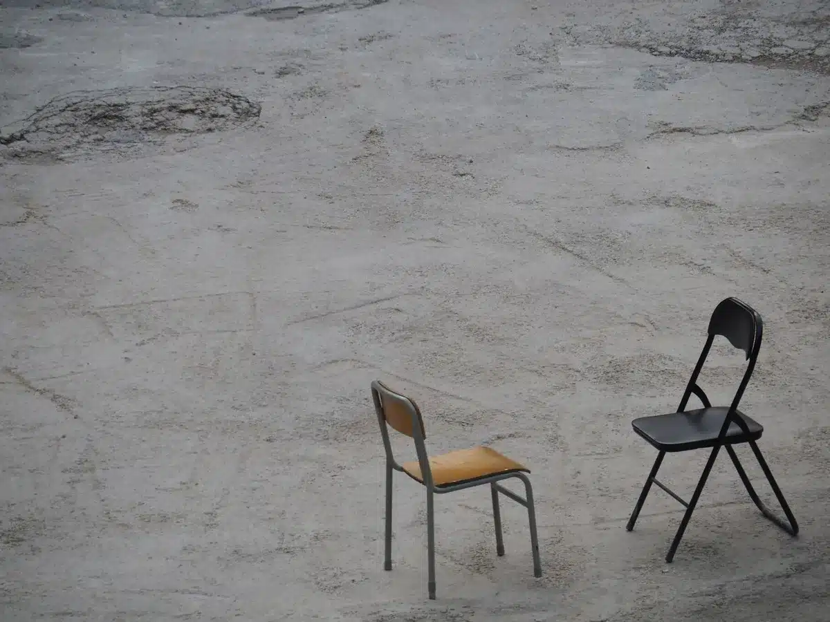 Two empty chairs facing each other in a dim room, a Bible on one table, a secular book on the other, a single dividing line of light down the center, symbolic contrast
