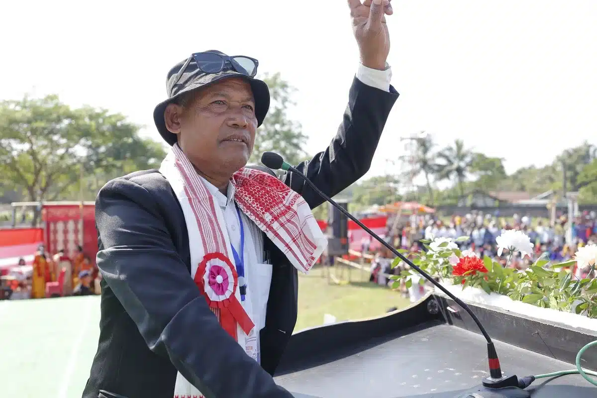 A politician at a podium with microphones during a campaign event, addressing supporters in a convention hall or rally setting, surrounded by campaign signage and American flags, conveying political ambition and public engagement