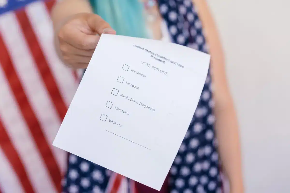 A voter at a polling place with a ballot, hand marking selections, with election officials and signage visible in the background