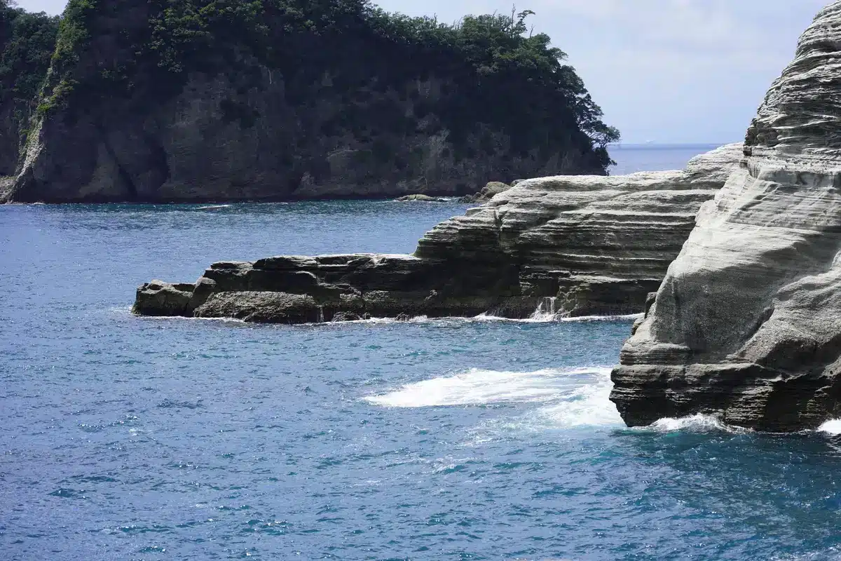 A wide view of a rocky volcanic coastline with crashing waves, steep cliffs rising from the ocean, no people visible, representing the Izu Islands' volcanic geology and seismic setting.