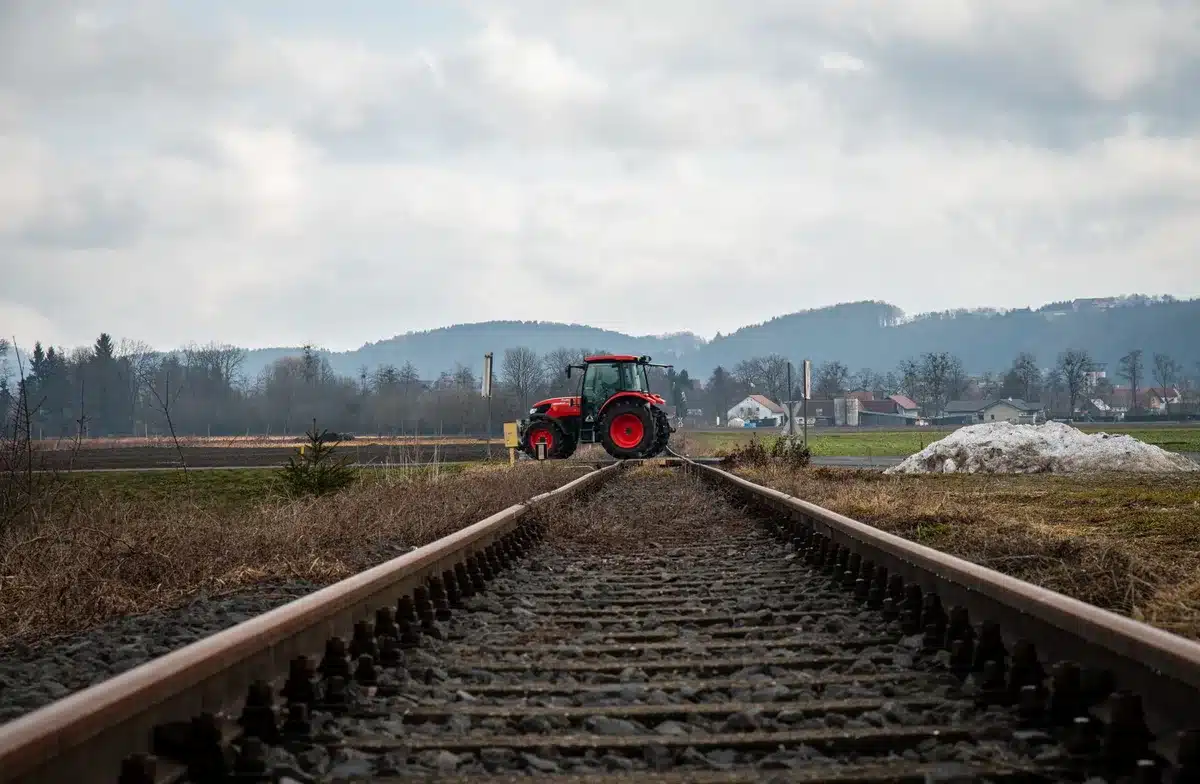 Freight train cars derailed and displaced from tracks in a wooded rural area, one locomotive tilted at an angle, surrounding forest visible, emergency response vehicles in the distance.