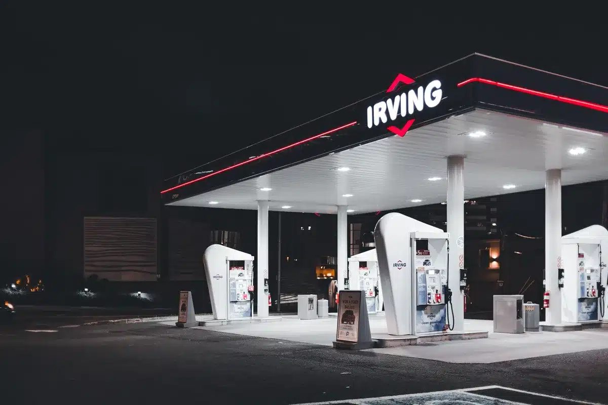 A gas pump nozzle at a station with a digital display showing a price above $4 per gallon, with blurred traffic and storefront lights in the background, evoking consumer anxiety.