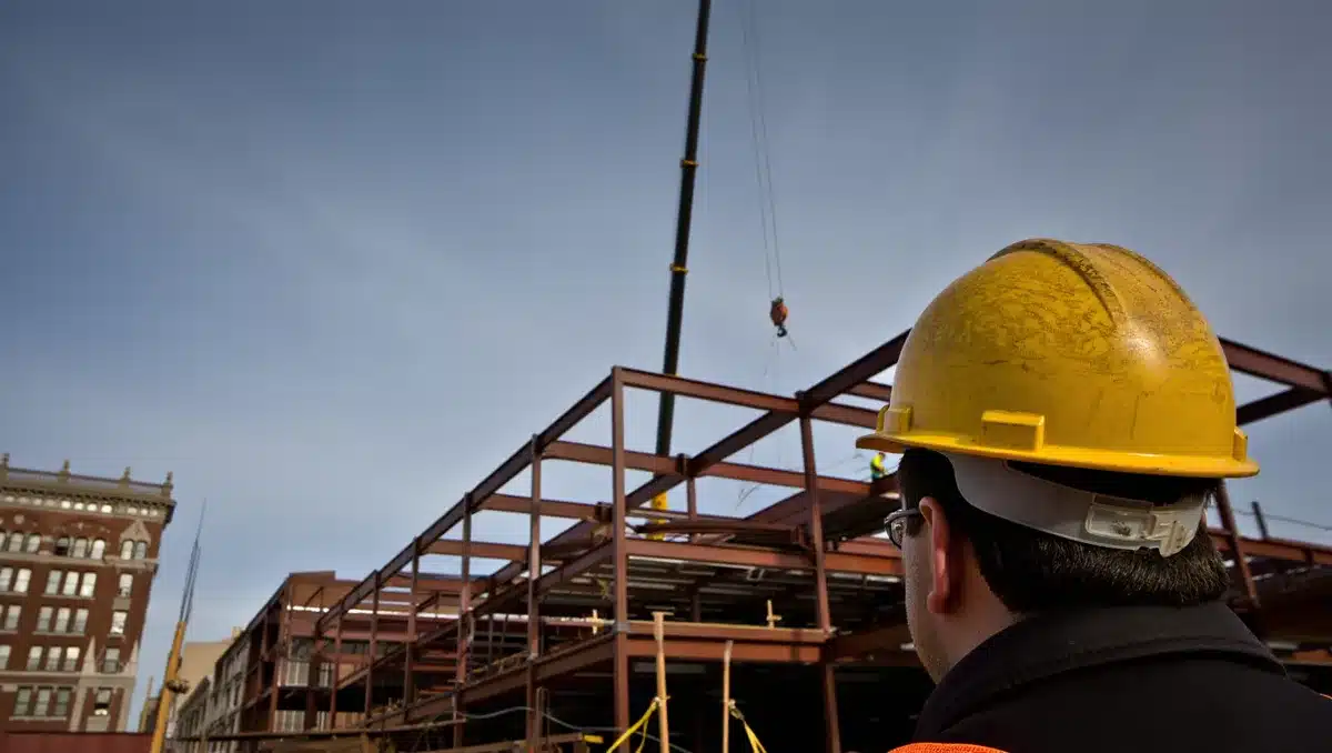 An anonymous figure in hard hat observing a large construction site with steel framing visible above ground, cranes overhead, and a historic building visible in the distance, tension.