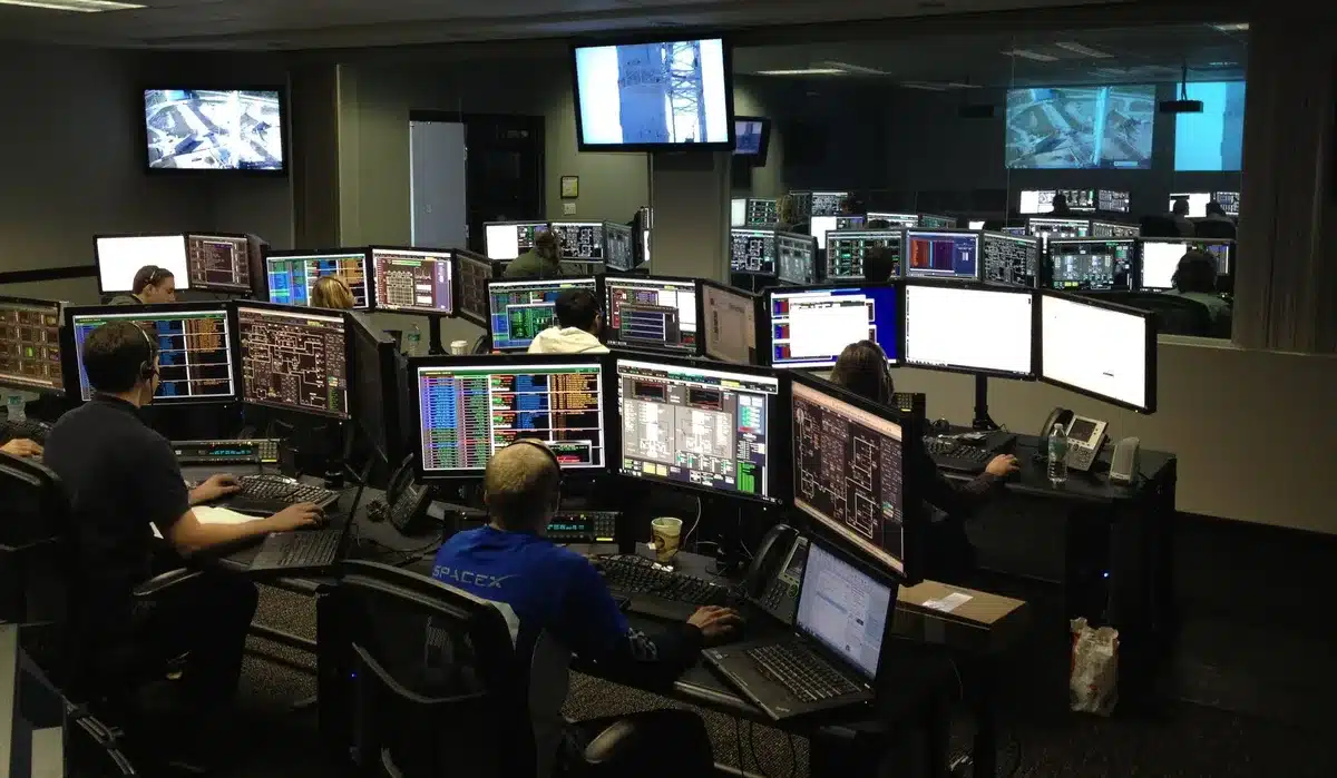 Military command center with glowing screens displaying tactical maps, officers in silhouette studying real-time strike data, tension visible in body language, dawn light through windows
