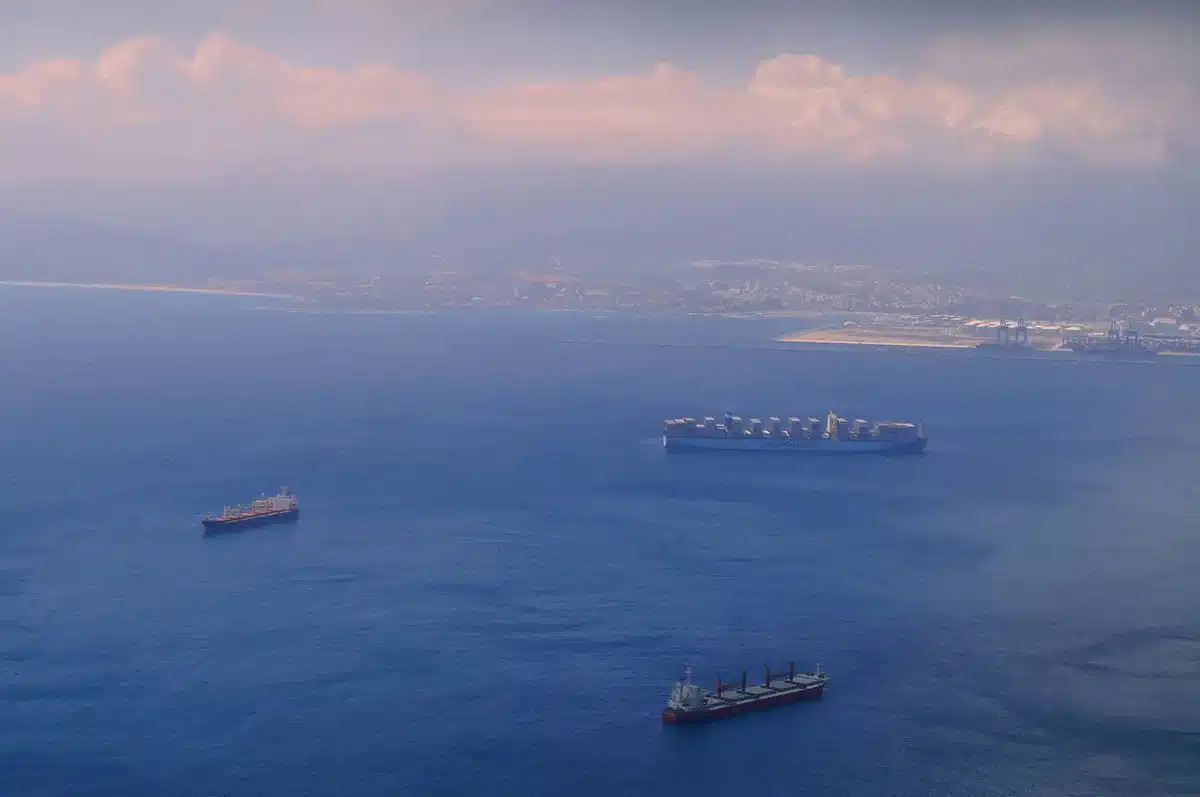 The Strait of Hormuz with a few cargo vessels anchored, calm waters, distant shoreline, GPS interference indicators on navigation screens