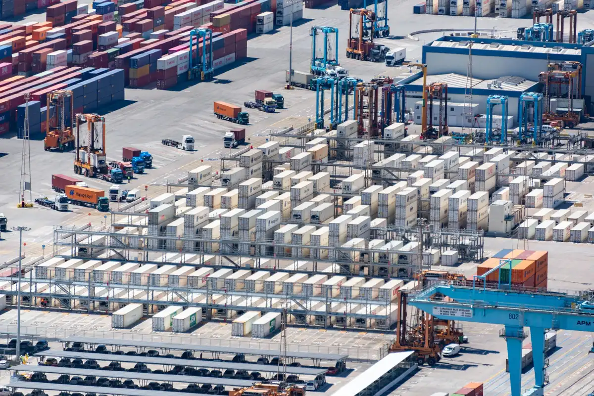 An empty cargo ship container terminal at dusk with shipping lanes visible in the distance, cranes silhouetted against the sky, the strategic waterway quiet but tense, a sense of economic pause