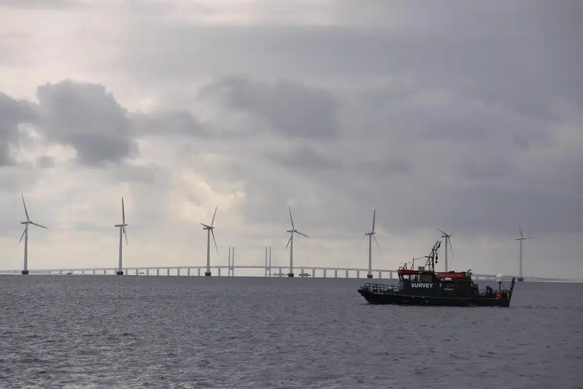 A sprawling offshore wind farm with turbines rising from blue-grey ocean waters under overcast skies, construction vessels nearby