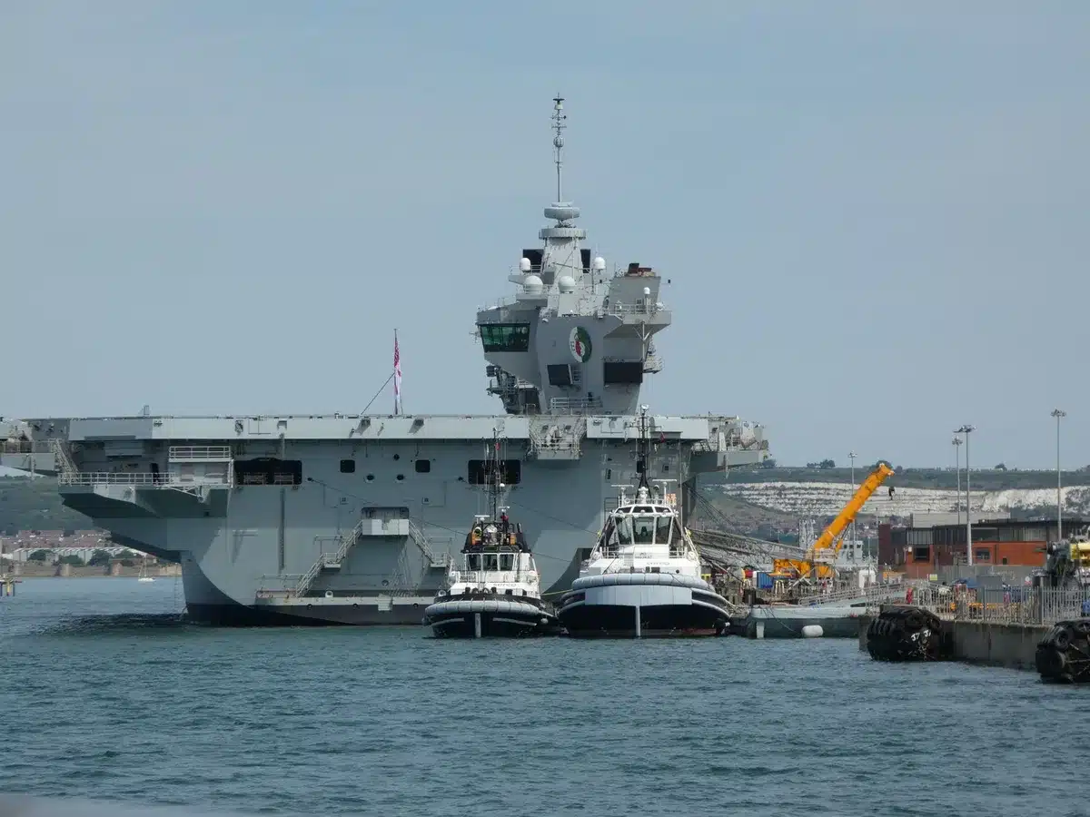 Military shipyard construction site with a large aircraft carrier hull under assembly, steel scaffolding and cranes visible, industrial activity and welding sparks, precision engineering