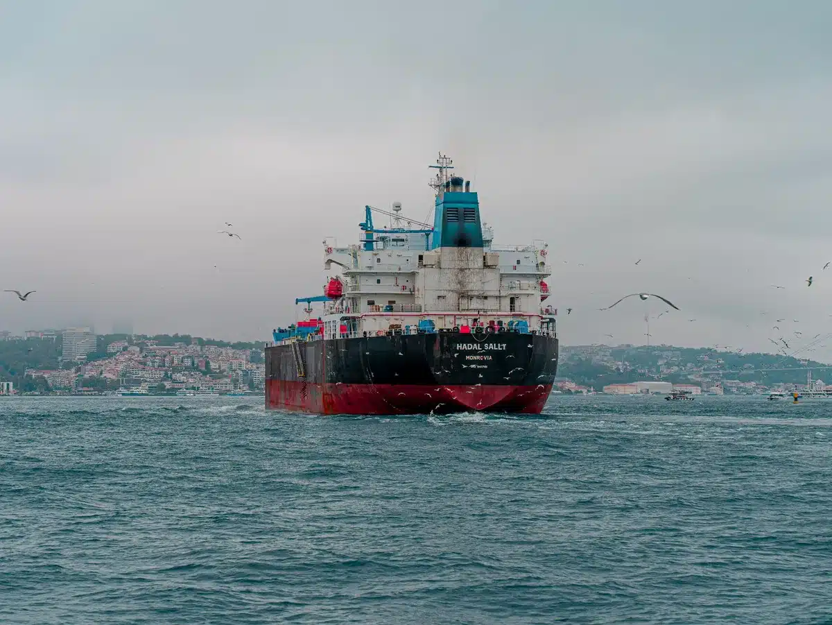 A naval blockade checkpoint with cargo vessels queued at a strategic waterway, military ships positioned along the channel, tension visible in the constrained shipping lanes