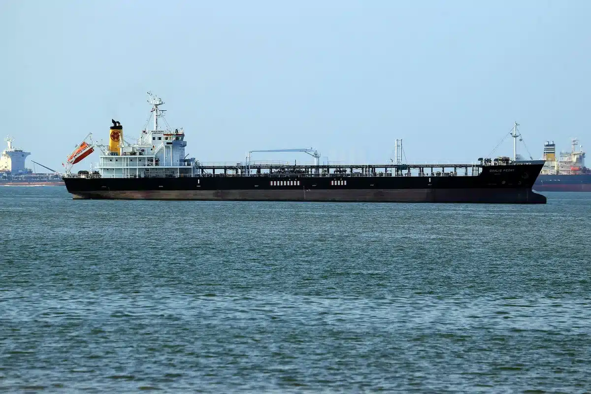 A cargo ship navigating through narrow straits with military vessels visible on the horizon, oil tankers stalled in queue, geopolitical tension evident in the constrained waterway