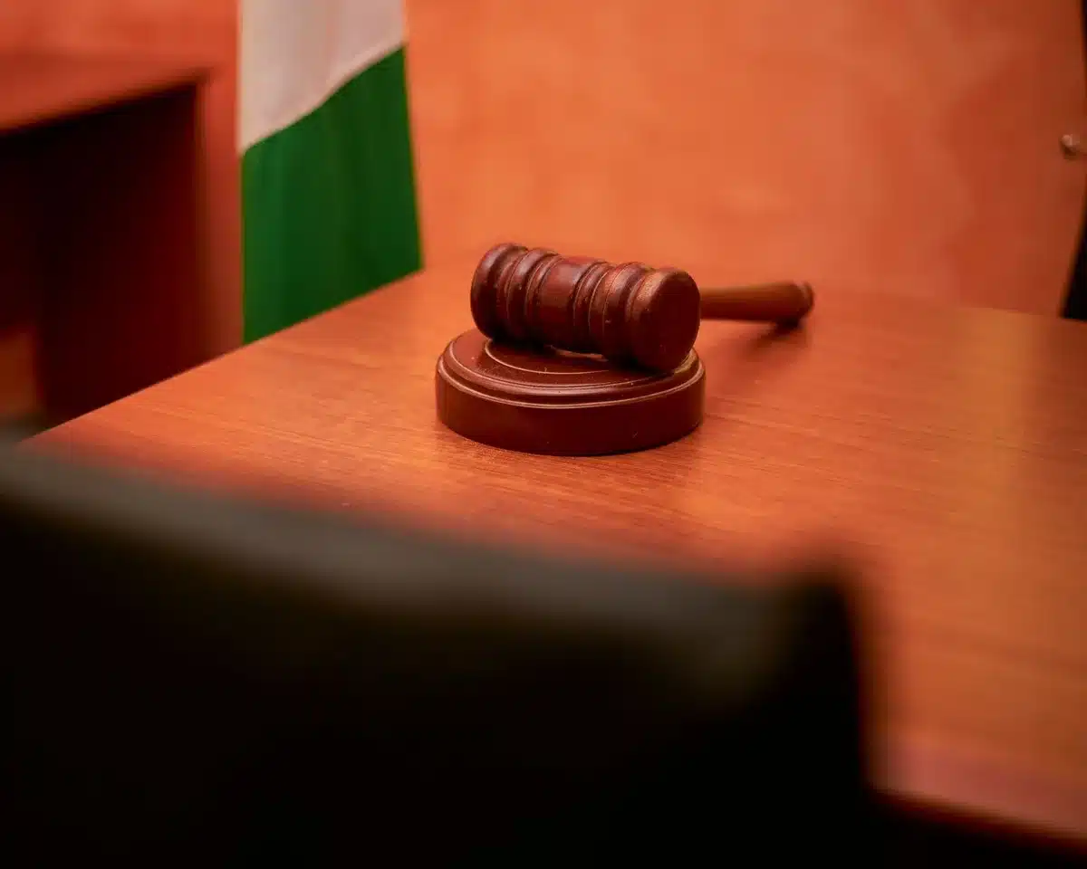A gavel resting on a wooden desk in a dim courtroom, with law books and a scales of justice in soft focus behind it, suggesting legal scrutiny