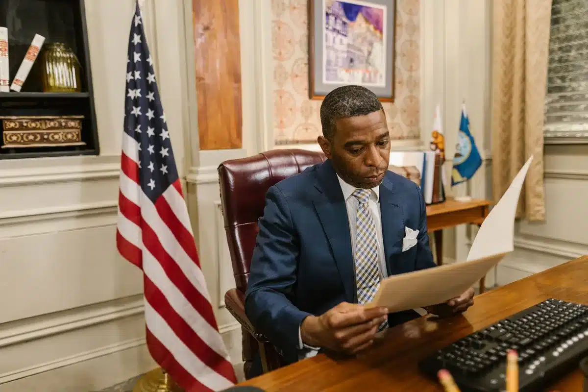An empty administrative desk with stacked documents and a nameplate, a window overlooking a distant national park landscape, tension in the austere office setting